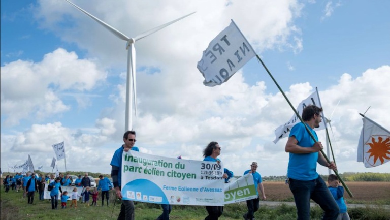 Un cortège d'hommes et de femmes, pour la plupart vêtus d'un tee-shirt bleu, défilent en bordure d'une route de campagne. Sur l'une des banderoles, on peut lire "Inauguration du parc éolien citoyen, Ferme éolienne d'Avessac". Derrière le cortège, une éolienne transperce l'horizon.