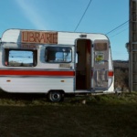 Une caravane blanche et rouge trône sur une pelouse en pleine campagne. Sur les rebords de ses fenêtres ouvertes est posé un carton où il est écrit en grosses lettres le mot 