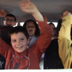 Un groupe d'enfants occupe tous les sièges de l'habitacle d'une voiture.Ils lèvent les bras en l'air et arborent des sourires radieux.