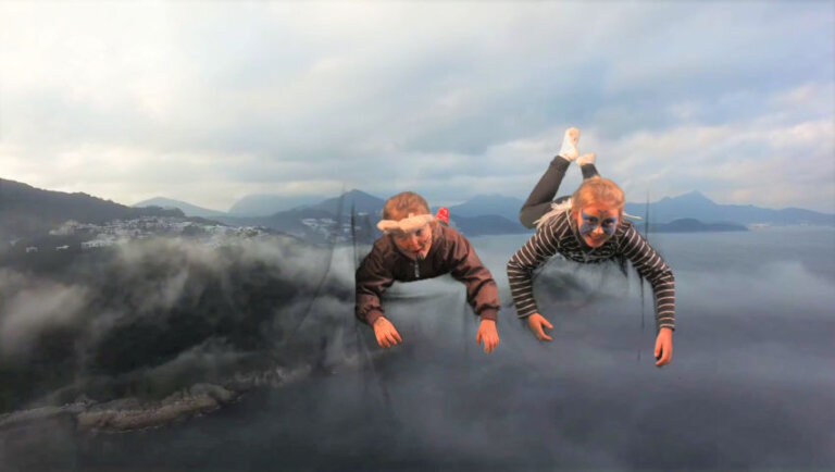 Deux enfants défuisés et maquillés semblent suspendus dans les airs, tout sourire. Derrière eux s'étend le large paysage des rives d'un grand lac entouré de montagnes embrumées.
