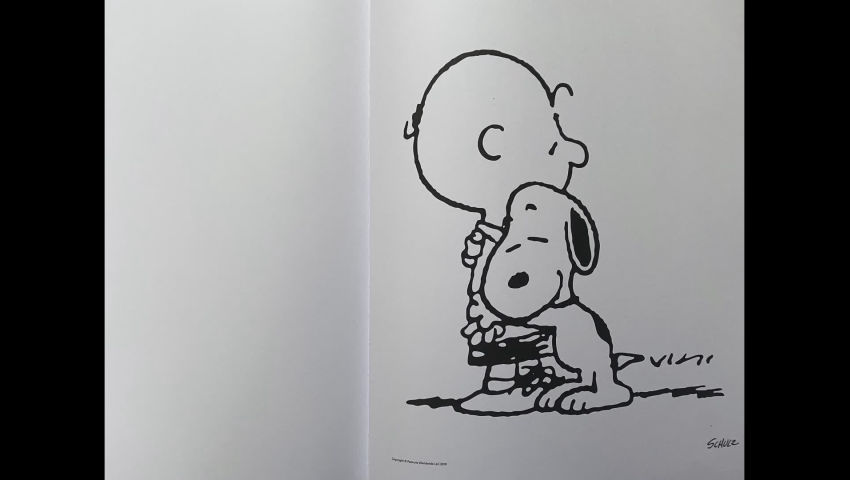 Sur la page d'un livre ouvert, un dessin signé par Schulz représente un enfant, qui reçoit un chalereux calin de la part de Snoopy, un chien bipède.
