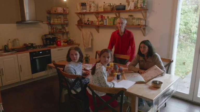 Une famille est réunie autour de la table de leur cuisine, où sont posés divers livres et cahiers. Ils tournent la tête vers l'extérieur de la pièce, la mine inquiète.